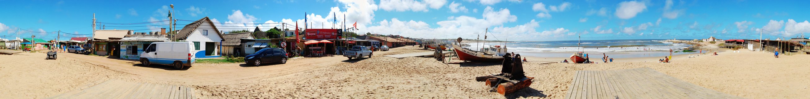 Wide panoramic view of a beach with fishing boats on sand, colorful beach houses, wooden boardwalks, and ocean waves under blue sky with clouds