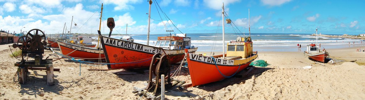 Multiple orange and red fishing boats with visible names Victoria II and Ana Carolina parked on sandy beach with ocean waves, blue sky with clouds, and rusty winch equipment in foreground