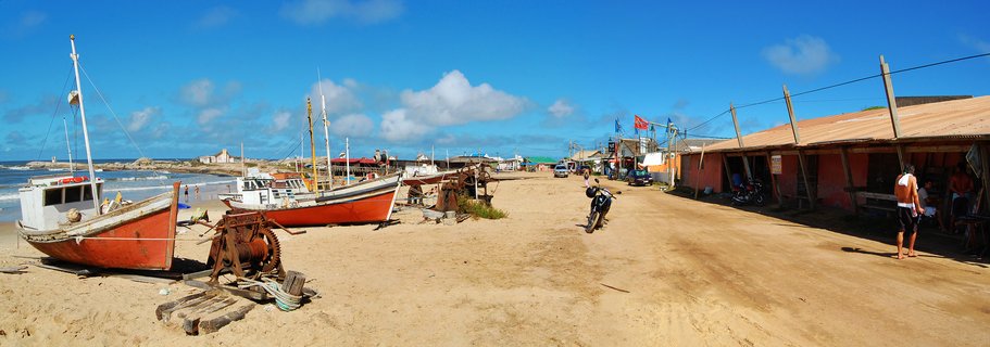 Two red fishing boats on a sandy beach with ocean waves on the left, rustic buildings on the right, blue sky with white clouds above, and a motorcycle parked on the sand