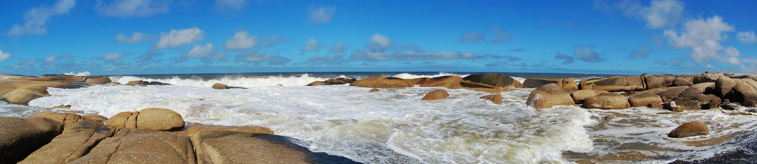 Ocean waves crashing against large tan boulders along a rocky coastline under a blue sky with scattered white clouds at Punta del Diablo, Uruguay
