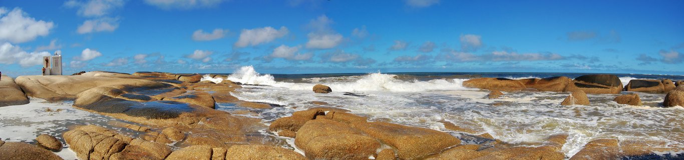 Panoramic view of golden-brown granite boulders on a rocky coastline with white ocean waves crashing between them, blue sky with scattered clouds above, and a small white building visible on the left side