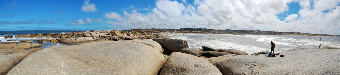 Wide coastal panorama showing large smooth granite boulders in foreground, ocean waves, a fisherman standing on rocks, and a small town on the horizon under blue sky with white clouds