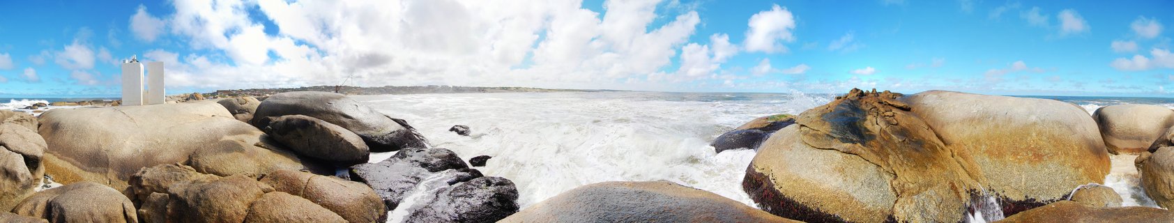 Panoramic view of large rounded granite boulders on a rocky coastline with white ocean waves crashing between them, blue sky with clouds above, and a white building visible on the left