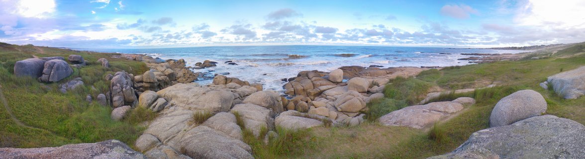 Panoramic view of large rounded granite boulders scattered across green coastal grassland leading to Atlantic Ocean waves breaking on rocky shore under partly cloudy blue sky
