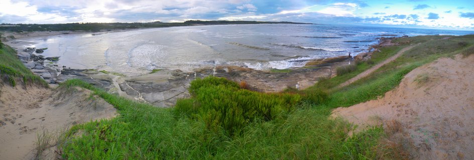 Panoramic coastal view from an elevated sand dune showing green vegetation in the foreground, rocky shoreline with layered formations, ocean waves, and cloudy sky over Punta del Diablo, Uruguay
