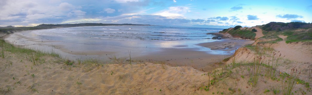Wide beach scene showing sand dunes with grass in the foreground, wet sand reflecting the sky, ocean waves, and cloudy sky over Punta del Diablo coastline
