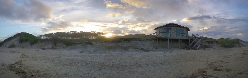 Wooden beach house on stilts among sand dunes at sunset with dramatic cloudy sky and tree line in Punta del Diablo, Uruguay