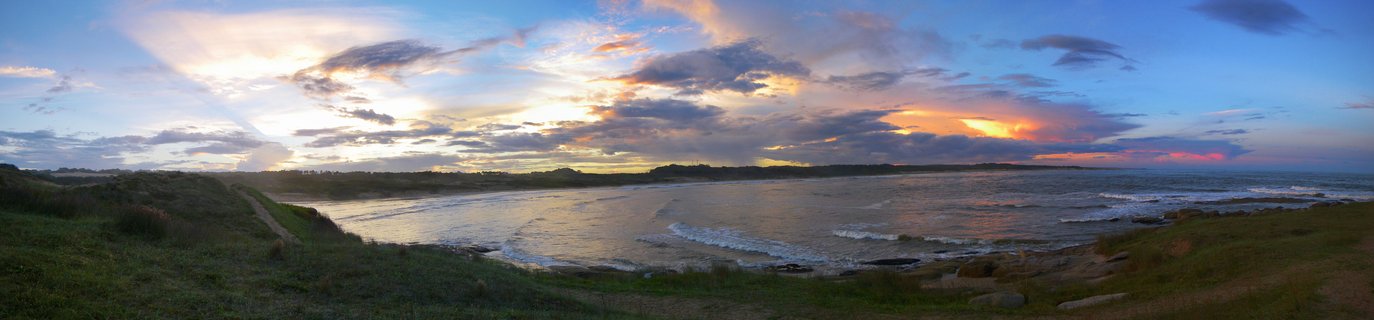 Panoramic view of sunset over a coastal bay with grassy dunes in foreground, waves rolling onto shore, and dramatic clouds with sun rays in the sky at Punta del Diablo, Uruguay