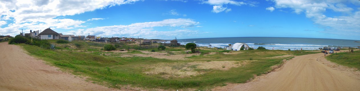 Wide coastal panorama showing two dirt roads leading through grassy dunes to Atlantic Ocean beach with small houses and blue sky with clouds in Punta del Diablo, Uruguay