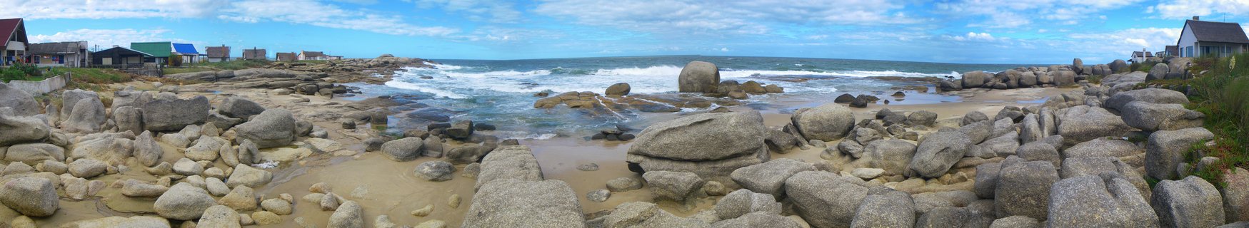 Wide coastal view of a rocky beach with large gray granite boulders on sand, ocean waves breaking in the middle distance, blue sky with white clouds, and small houses visible along the shoreline