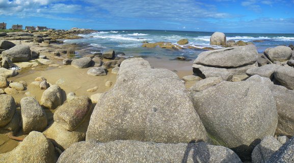 Large gray granite boulders scattered across a sandy beach with ocean waves and blue sky with clouds at Punta del Diablo, Uruguay