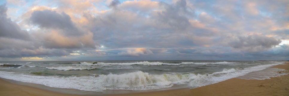 Panoramic view of ocean waves breaking on a sandy beach at Punta del Diablo, Uruguay, with dramatic cloudy sky at sunset showing pink and orange highlights