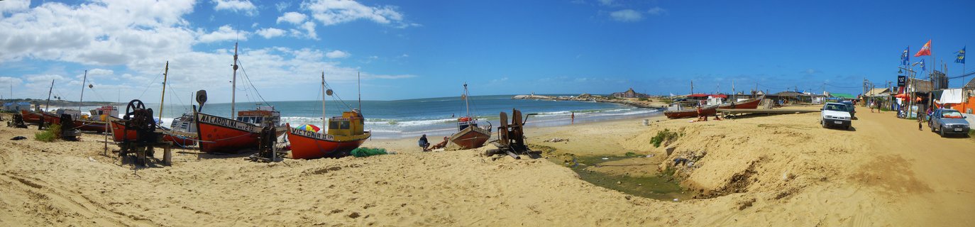 Panoramic view of colorful fishing boats pulled up on sandy beach with ocean waves, blue sky with clouds, and coastal village buildings in Punta del Diablo, Uruguay