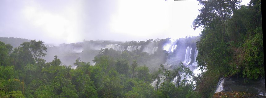 Wide panoramic view of Iguazu Falls showing multiple waterfall cascades surrounded by dense green tropical forest with heavy mist rising from the water