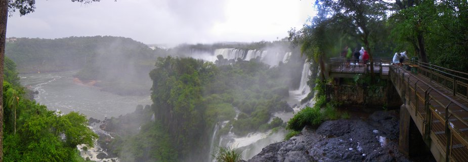 Panoramic view of Iguazu Falls showing multiple waterfall cascades surrounded by lush green rainforest, with tourists standing on an observation walkway on the right side