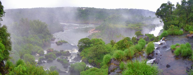 Wide panoramic view of Iguazu Falls with multiple water cascades flowing through lush green tropical vegetation with mist rising from the river below