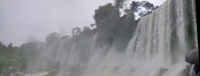 Multiple powerful waterfall cascades at Iguazu Falls with heavy mist, tropical green trees on cliff tops, and a person holding an umbrella visible on the right side