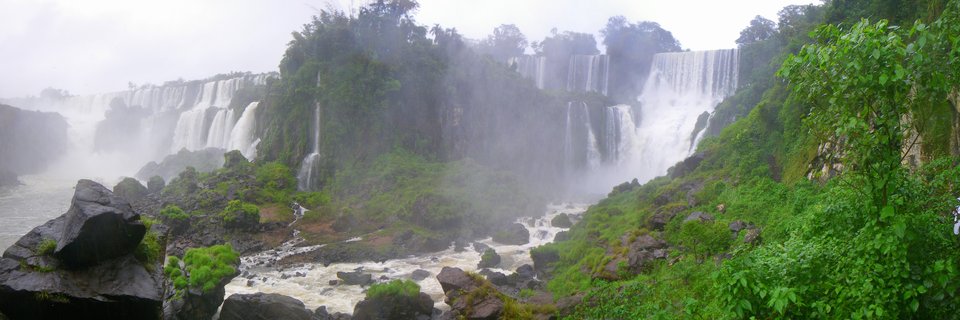 Wide panoramic photograph of Iguazu Falls showing multiple waterfall cascades flowing over rocky cliffs surrounded by dense green tropical vegetation with mist rising from the base