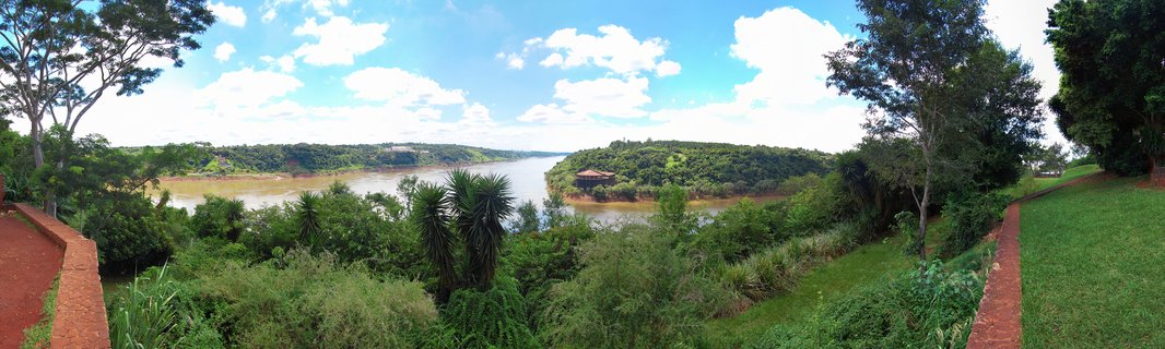 Wide panoramic landscape showing the Iguazu River flowing through dense green tropical forest with red brick viewing platform edges visible on both sides, blue sky with white clouds above