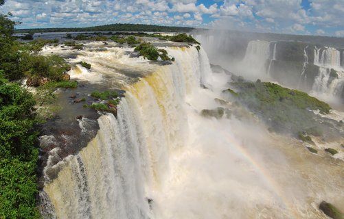 Multiple powerful waterfalls cascading over rocky cliffs with lush green vegetation, rainbow visible in mist, blue sky with white clouds above