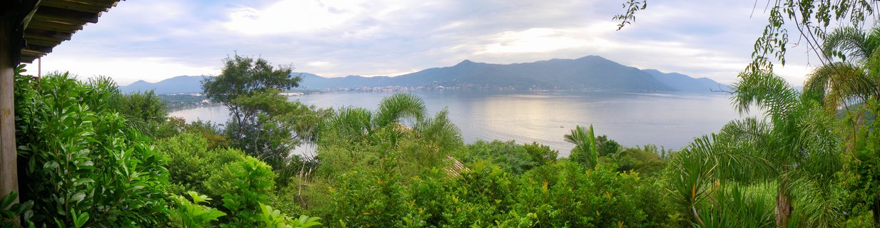 Panoramic coastal landscape showing tropical green vegetation in foreground, calm bay waters in middle ground, and mountain ranges in background under cloudy sky