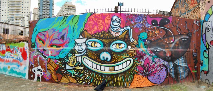 Colorful street art mural painted on a wall featuring a large cat face with bright green eyes, white teeth grin, surrounded by psychedelic patterns, cartoon sailor character, and vibrant graffiti elements with tall buildings in background