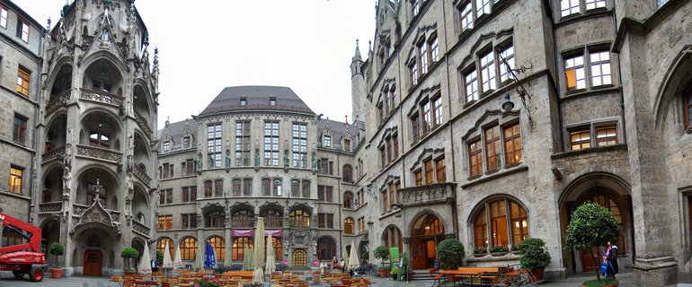 Stone courtyard surrounded by Gothic Revival buildings with ornate facades, arched windows, decorative sculptures, outdoor wooden tables and chairs, potted trees, Munich Germany