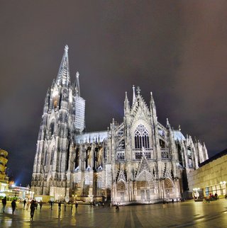 Night photograph of Cologne Cathedral showing illuminated Gothic twin spires, detailed stone facade with pointed arches and stained glass windows, people walking on wet paved plaza in foreground