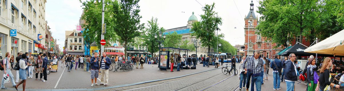 Wide street scene in Amsterdam showing numerous pedestrians walking on cobblestone pavement with tram tracks, historic buildings including a red-brick tower structure, green trees, bicycle parking, and outdoor cafe seating