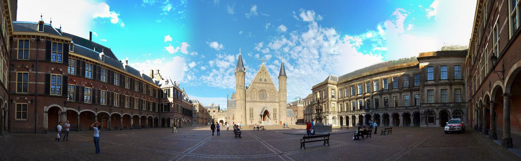 Wide courtyard view of the Binnenhof in The Hague showing the Gothic Ridderzaal Hall of Knights flanked by historic brick buildings with arched walkways under a blue sky with scattered clouds