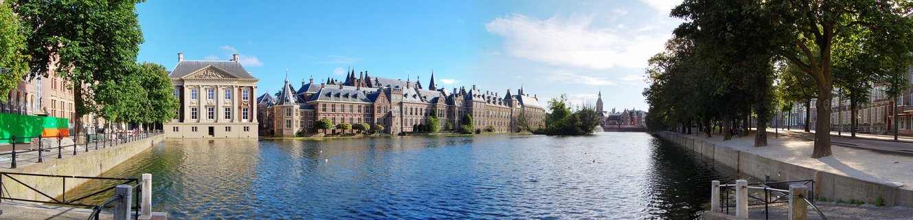 Panoramic view of historic parliament buildings with Gothic spires and towers along a canal with tree-lined walkways on both sides under blue sky