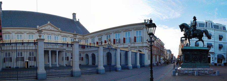 Ornate black and gold palace gates with stone pillars in front of a classical Dutch palace building, with a bronze equestrian statue on a stone pedestal in the foreground on a cobblestone plaza