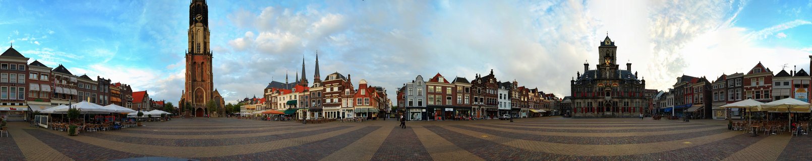 Wide panoramic street view of a Dutch town square with a tall brick church tower on the left, historic buildings with gabled roofs, outdoor café terraces with white umbrellas, and an ornate town hall building on the right under a blue sky with scattered clouds