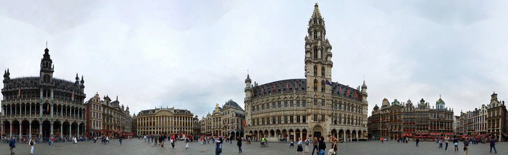 Panoramic view of Grand Place square in Brussels showing Gothic Town Hall tower and ornate historic buildings with people walking on cobblestone plaza under cloudy sky