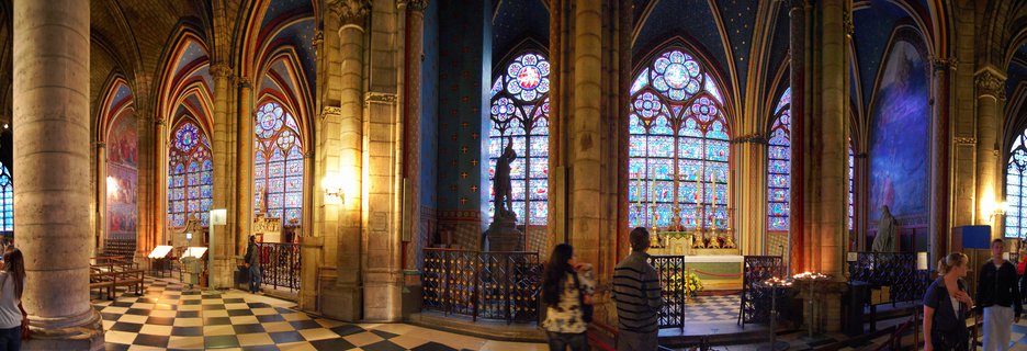 Interior view of Sainte-Chapelle showing Gothic stone columns, pointed arches, colorful medieval stained glass windows, blue walls with gold stars, and visitors on a checkered floor