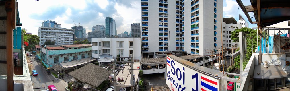 Elevated street view of Bangkok showing white apartment buildings with blue awnings, a guesthouse sign with Thai flag colors, and modern skyscrapers in the background under cloudy sky
