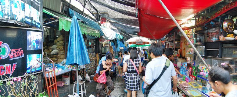 People walking through a covered outdoor market alley in Bangkok with vendor stalls on both sides under colorful tarps