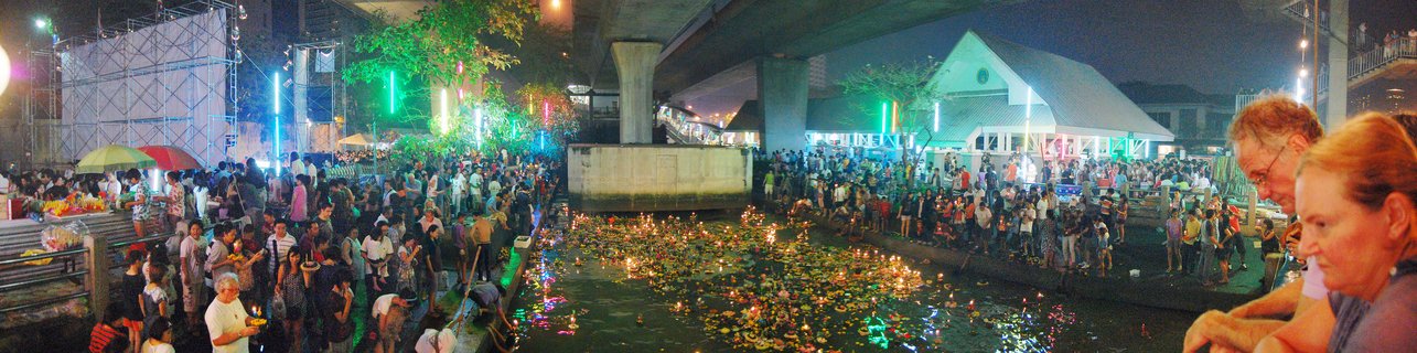 Night festival scene showing crowds of people along both sides of a canal filled with floating lit lanterns, with an elevated concrete structure overhead and colorful stage lighting