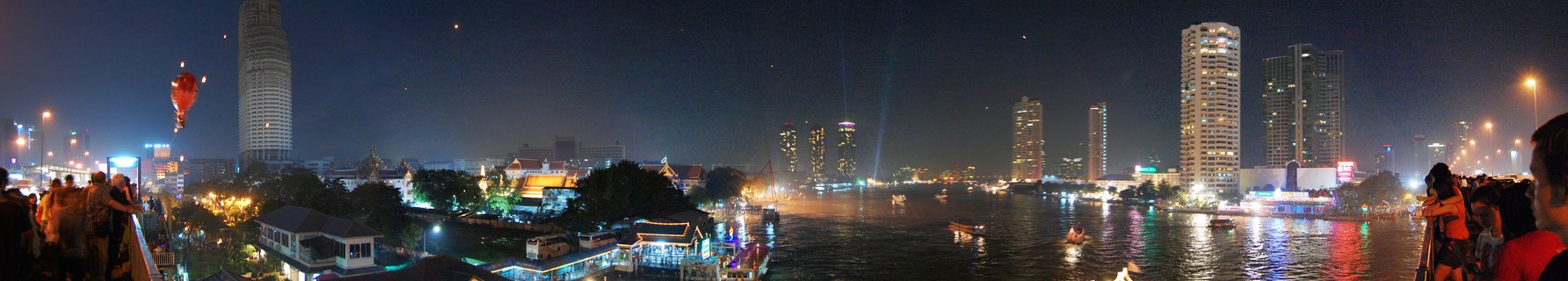 Nighttime panoramic view of Bangkok's Chao Phraya River with illuminated skyscrapers, boats on the water, and people viewing from riverside platforms