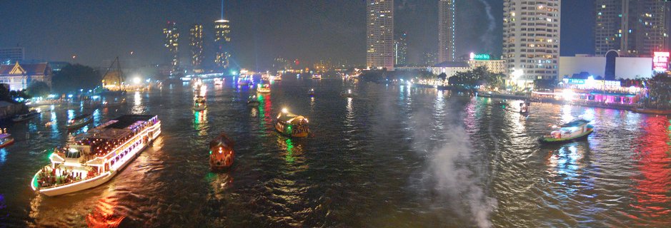 Night photograph of multiple illuminated boats on a wide river with Bangkok city skyline featuring tall buildings and colorful light reflections on water