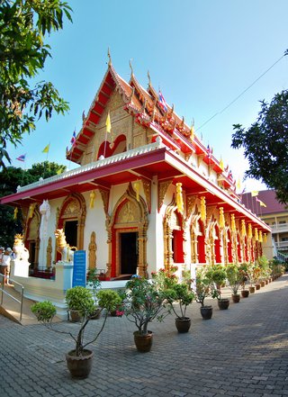 Thai Buddhist temple with ornate golden decorations, red roof tiles, guardian lion statues, and potted plants in courtyard under clear blue sky