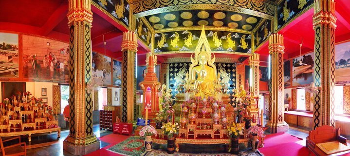 Golden Buddha statue in meditation pose inside Thai Buddhist temple with ornate red and gold ceiling, decorative columns, flower offerings, and traditional wall murals