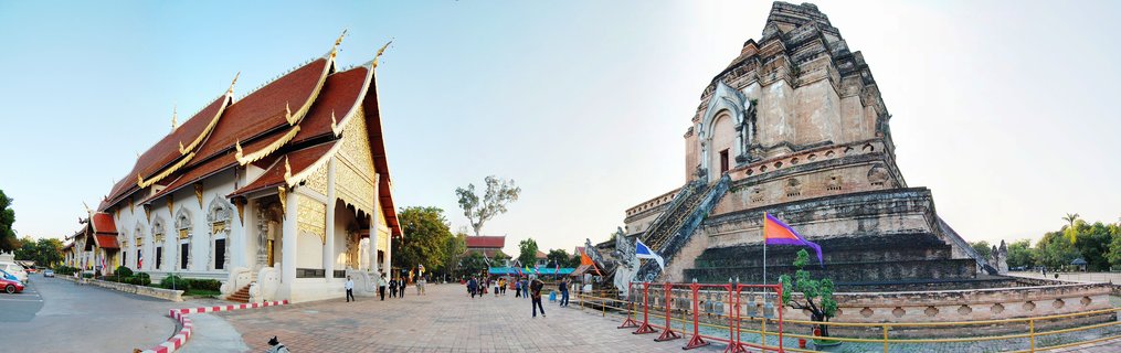 Panoramic view of Wat Chedi Luang Buddhist temple showing large ancient brick chedi stupa on right and white temple building with ornate red and gold roof on left, with visitors walking on courtyard between structures