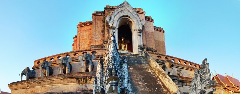 Large brick Buddhist temple chedi with stone elephant statues along the base, golden Buddha statue in an arched niche, and ornate naga serpent sculptures in the foreground against blue sky