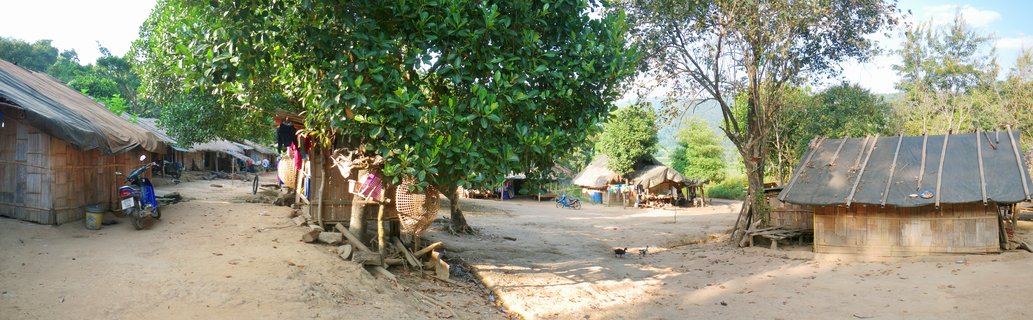 Rural village scene showing traditional bamboo huts with metal roofs along a dirt road, large green trees, a blue motorcycle, and mountains visible in the background under a partly cloudy sky