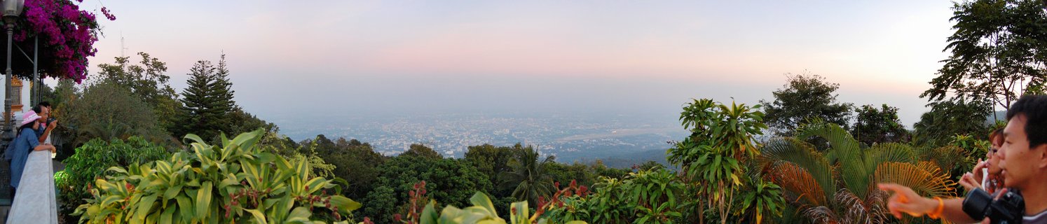 Panoramic view from Doi Suthep temple showing tourists at a stone railing overlooking Chiang Mai city through tropical vegetation at dusk with pink and blue sky