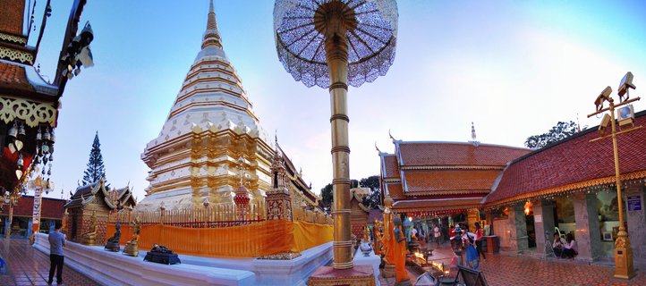 White and gold Buddhist stupa at Thai temple complex with monks in orange robes, ornate golden umbrella canopy, traditional temple buildings with red tile roofs, and visitors in courtyard