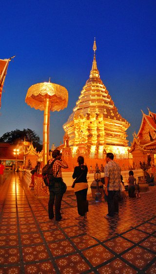 Illuminated golden Buddhist temple chedi at twilight with visitors on ornate tiled terrace, Chiang Mai, Thailand