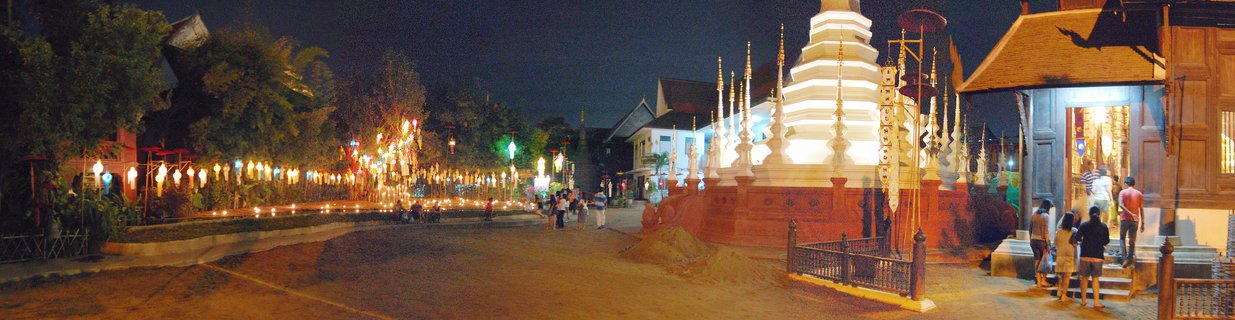 Night view of an illuminated white Buddhist stupa with tiered levels at a temple in Chiang Mai, Thailand, with strings of glowing lanterns and visitors walking on temple grounds