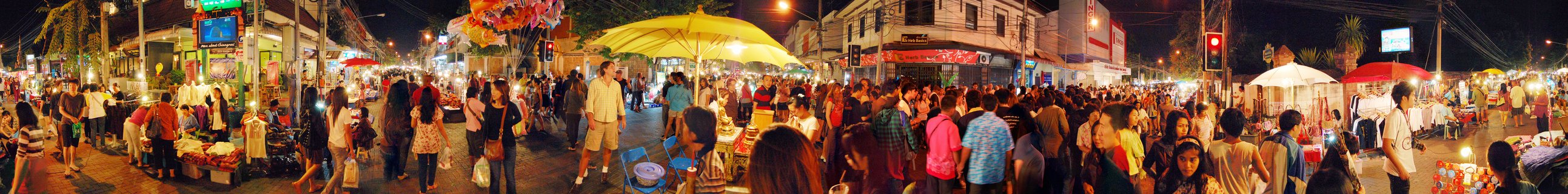 A crowded pedestrian street at night in Chiang Mai filled with people browsing vendor stalls under yellow white and red umbrellas with shops and buildings lining both sides